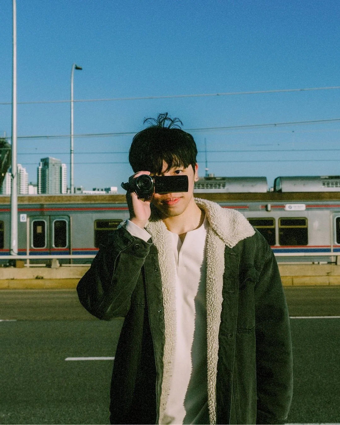 A young man with black hair is holding a video camera up to his eye. He is standing outdoors on a street in front of a train and city skyline, wearing a black jacket with a white shearling collar over a white shirt under a clear sky.