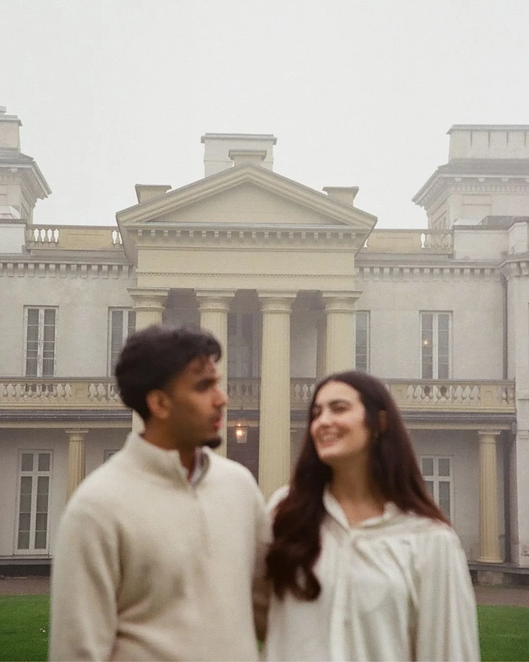 A young man and woman standing outside in front of a large, white, colonial-style mansion with columns and balconies, on a foggy day.