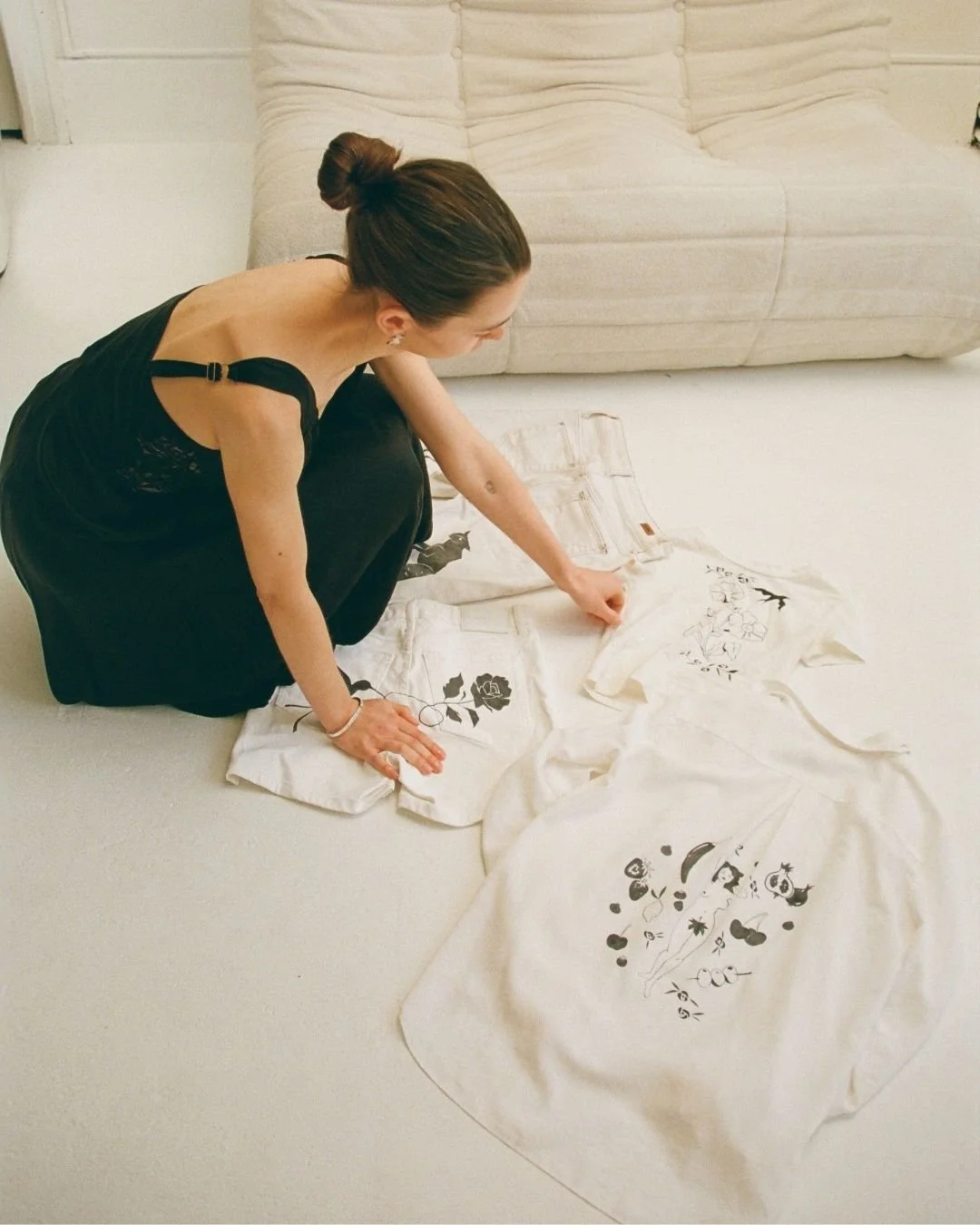 A woman with dark hair in a bun, wearing a black dress, sits on the floor and arranges white clothes with black floral and abstract designs.