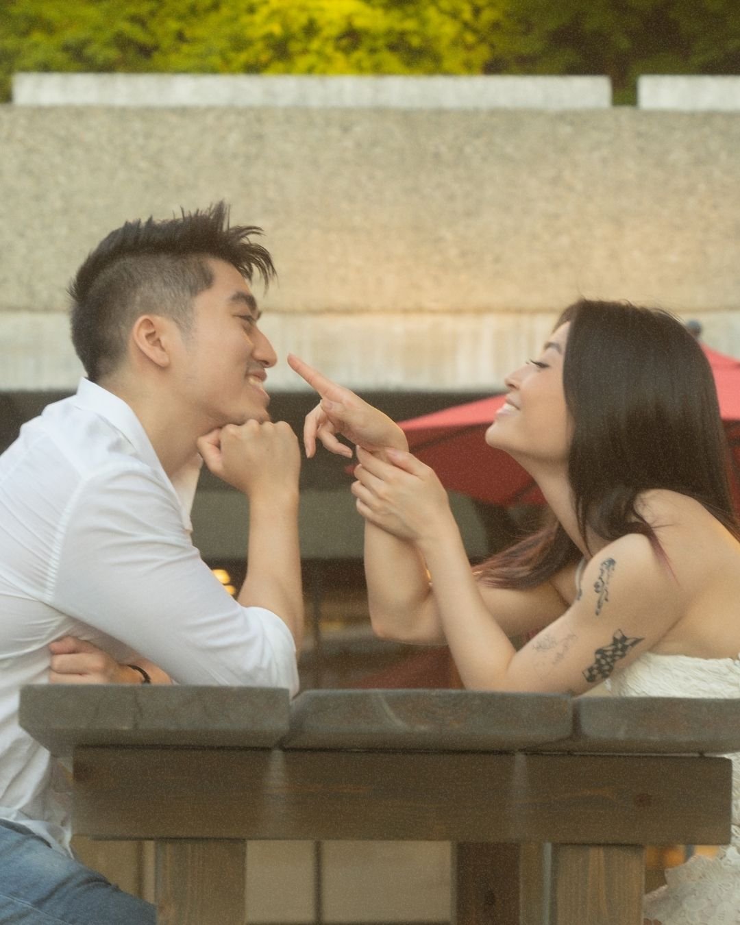 A young man and woman smiling and playfully interacting at an outdoor table, with a red umbrella in the background.