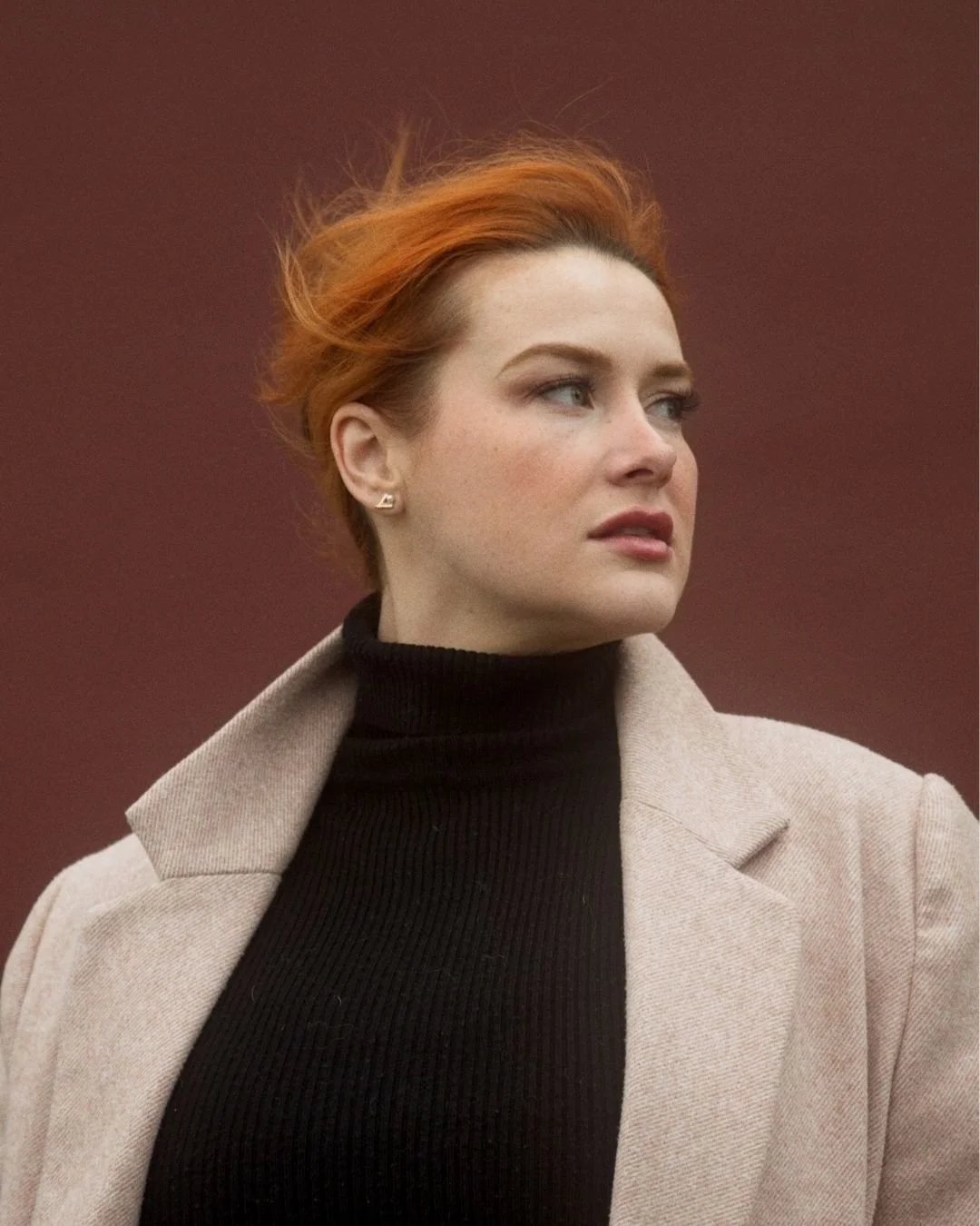 A woman with short red hair wearing a beige coat over a black turtleneck sweater, standing outdoors against a blurred background.