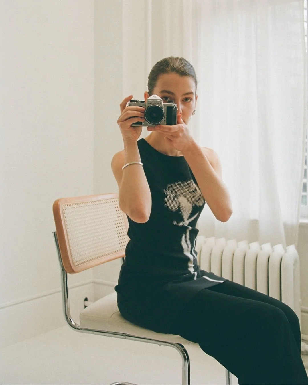 A woman sitting on a chair taking a selfie with a vintage camera in a bright room with white walls, sheer curtains, and a radiator.