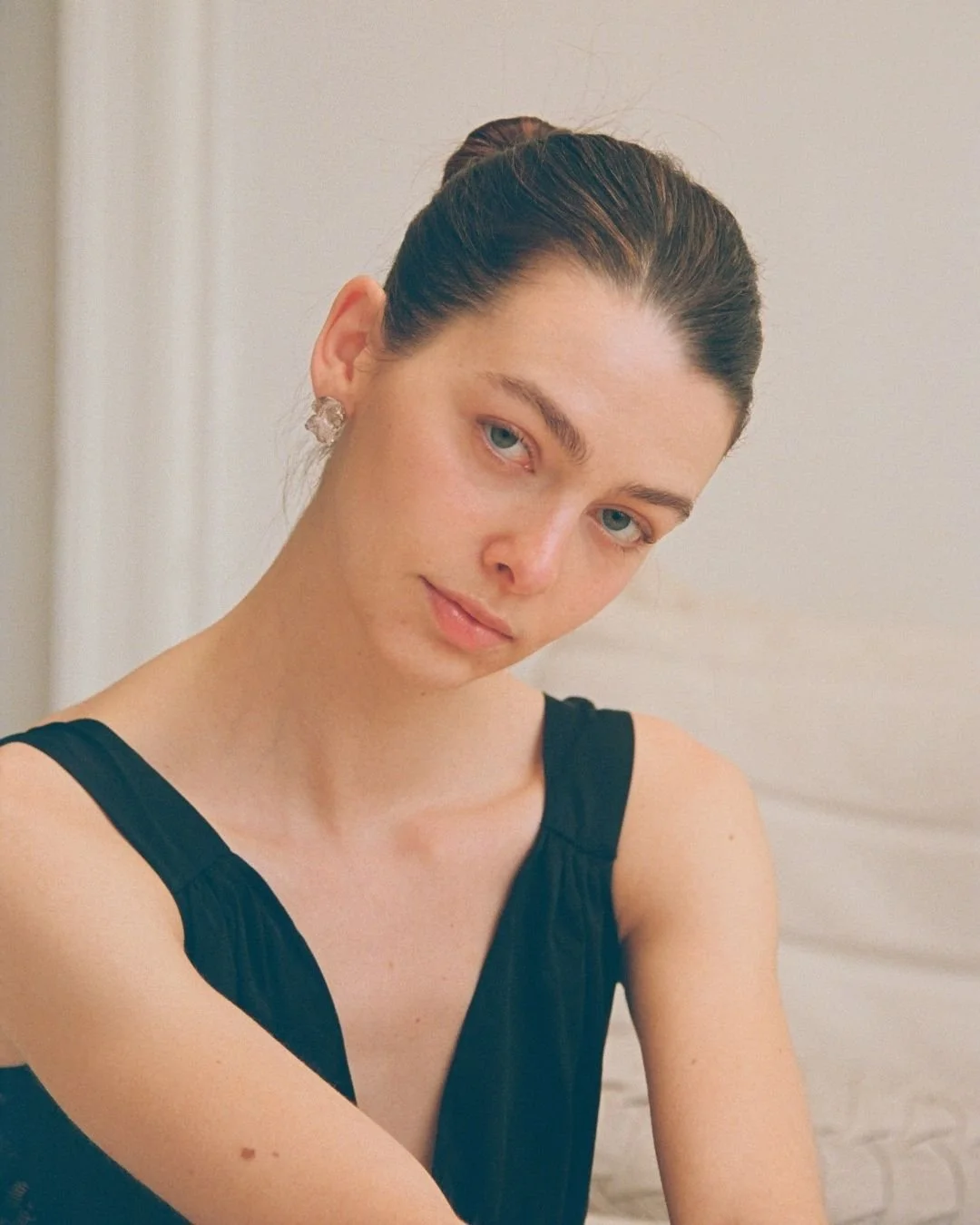 A young woman with light skin and blue eyes, wearing a black sleeveless top, silver earrings, and a tai l. Her hair is styled in a bun, and she is looking slightly toward the camera with a neutral expression, against a neutral background.