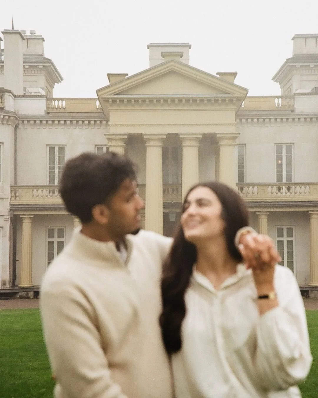 A young man and woman smiling and looking at each other outdoors in front of a large, classical mansion with columns and balconies.
