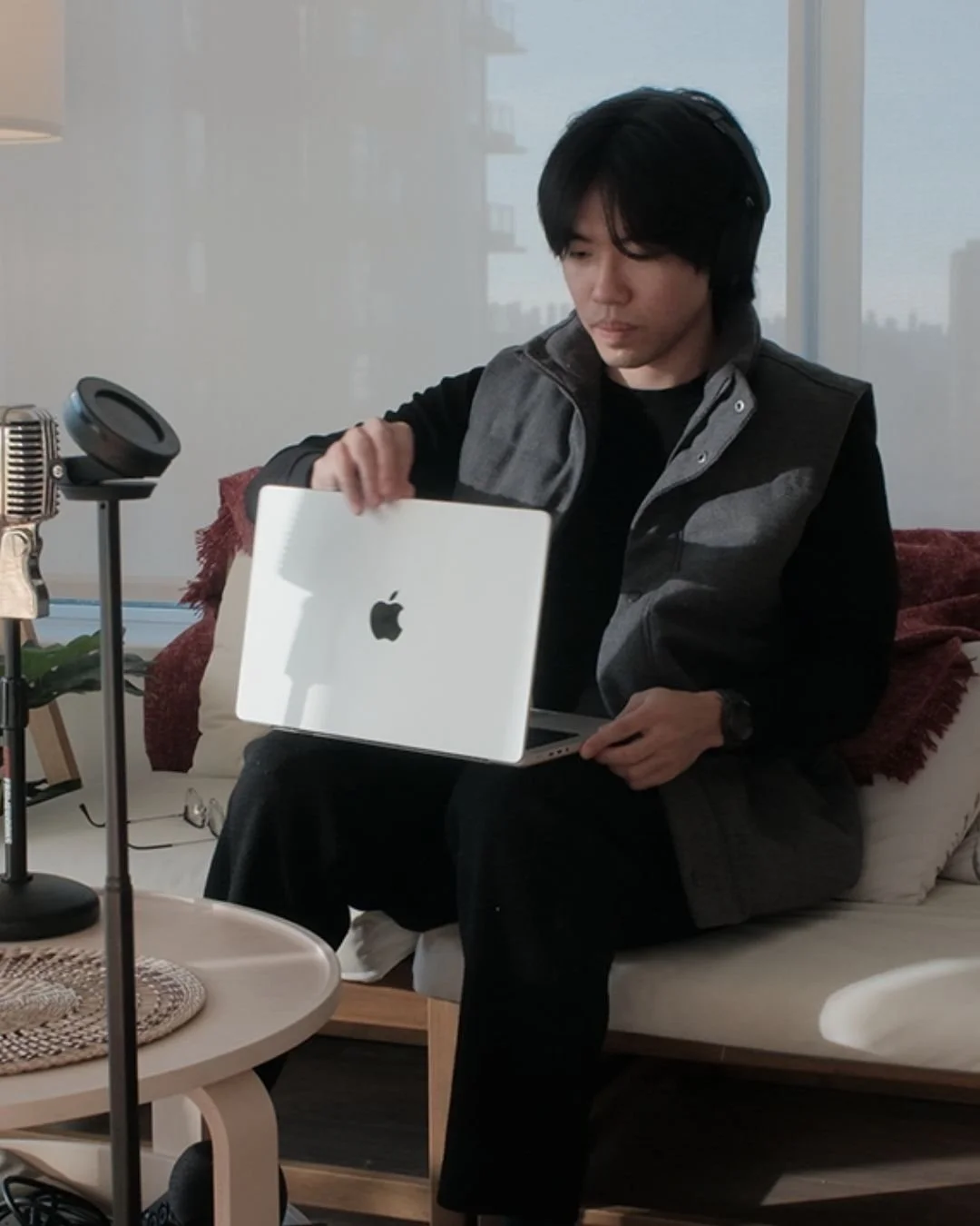 A man sitting on a couch holding an open Apple MacBook, with a vintage microphone and a recording device on a stand nearby, in a room with a large window.