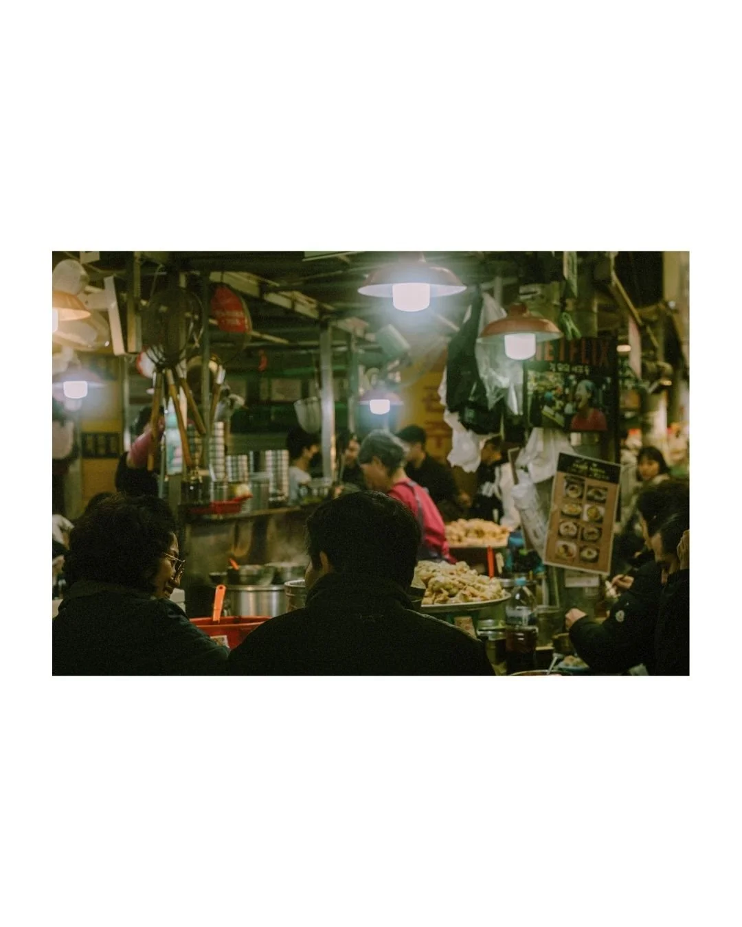 A busy indoor restaurant or food stall with customers eating and staff preparing food, featuring hanging lights and various kitchen utensils.