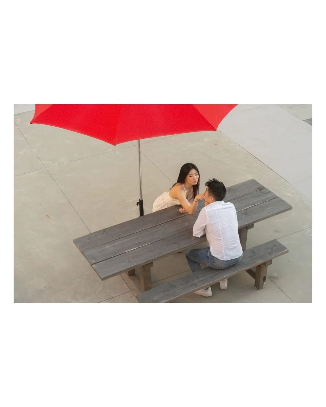 A young woman and man sitting at a wooden picnic table under a large red umbrella on a concrete surface in Simon Fraser University, Burnaby B.C.