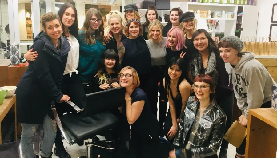 Group of women and girls smiling and gathered together indoors, some making peace signs, with shelves and decorative items in the background.