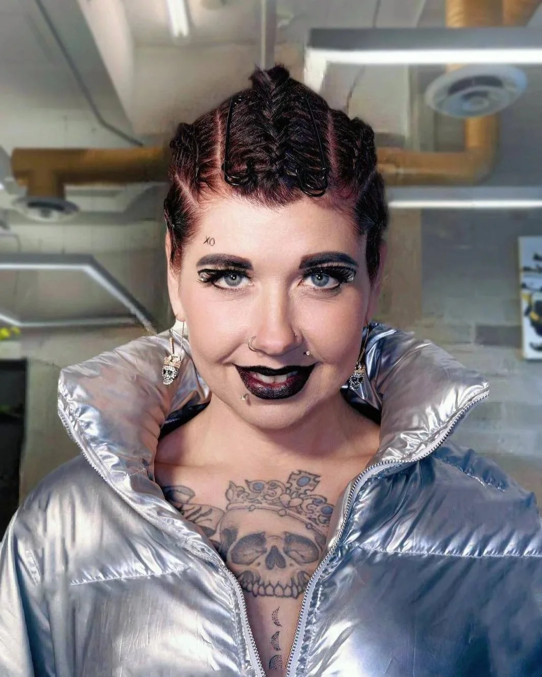 A woman with short curly dark hair styled with hair clips, wearing makeup with dark lipstick, earrings, and tattoos on her chest. She is dressed in a shiny silver jacket, smiling in an industrial setting.