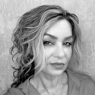 A woman with wavy hair and makeup looking at the camera, wearing a shirt and a necklace.