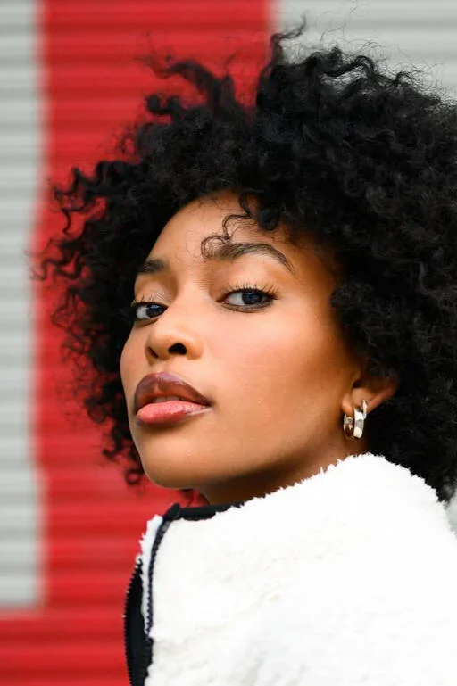 Close-up of a woman with curly black hair, wearing earrings, looking over her shoulder against a background with red, gray, and white stripes.