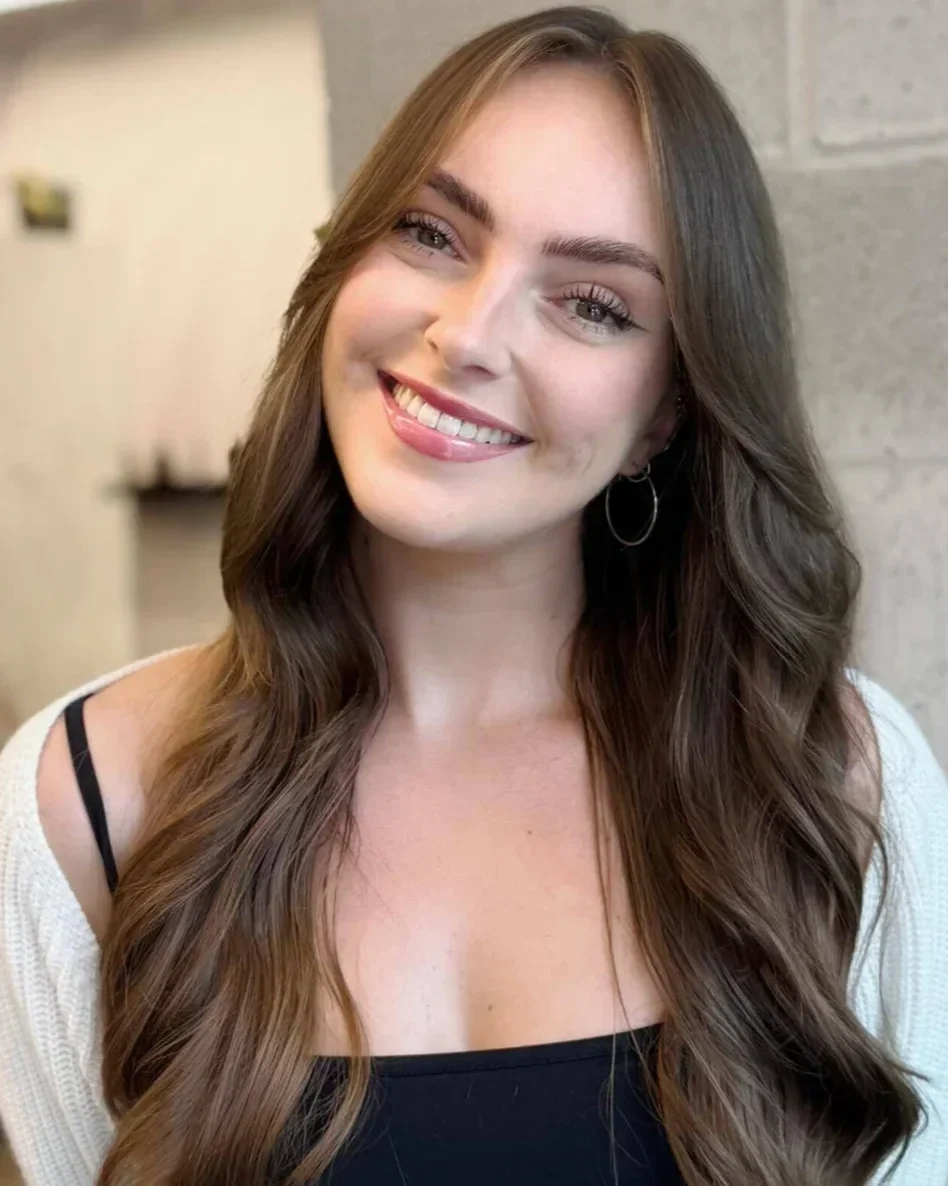 A young woman with long, wavy brown hair, smiling, wearing a black top and a cream cardigan, with hoop earrings, standing in front of a beige wall.
