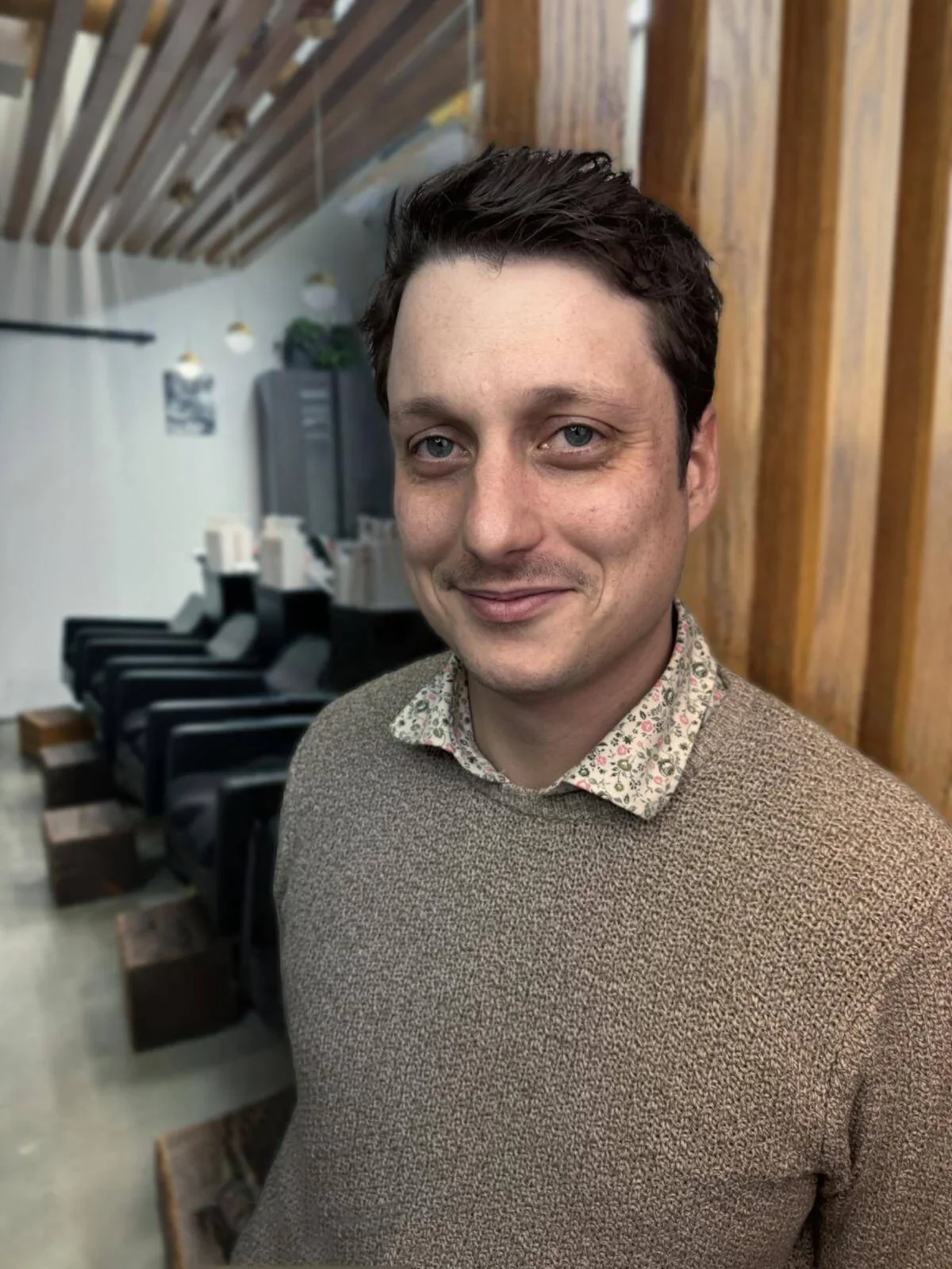 A man with dark hair and blue eyes smiling, wearing a beige sweater over a floral collared shirt, standing inside a room with wooden walls and black chairs in the background.
