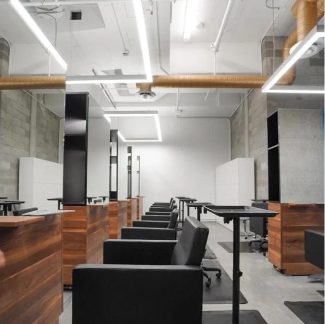 Empty modern salon with black chairs and workstations separated by wooden dividers, white walls, and industrial ceiling with exposed pipes and linear LED lighting