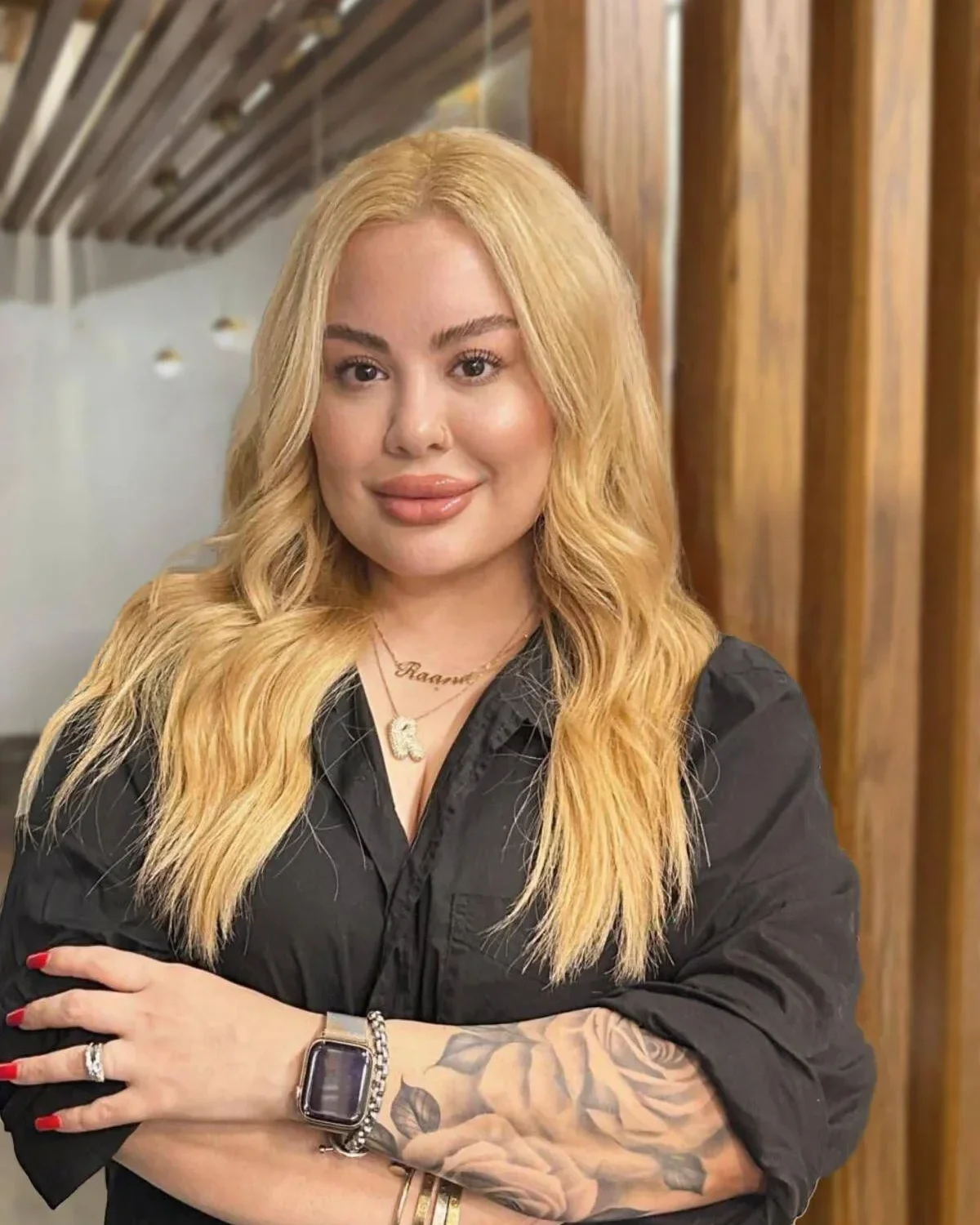 A woman with long blonde hair, wearing a black shirt, gold jewelry, a watch, and a tattooed arm, standing indoors in front of wooden paneling.