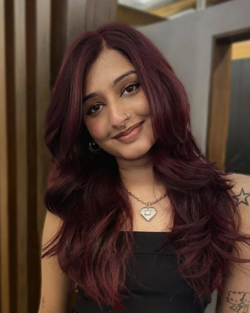 A woman with shoulder-length maroon hair smiling at the camera, wearing a black top, a heart-shaped necklace, and a nose ring, with tattoos on her left arm, in an indoor setting.