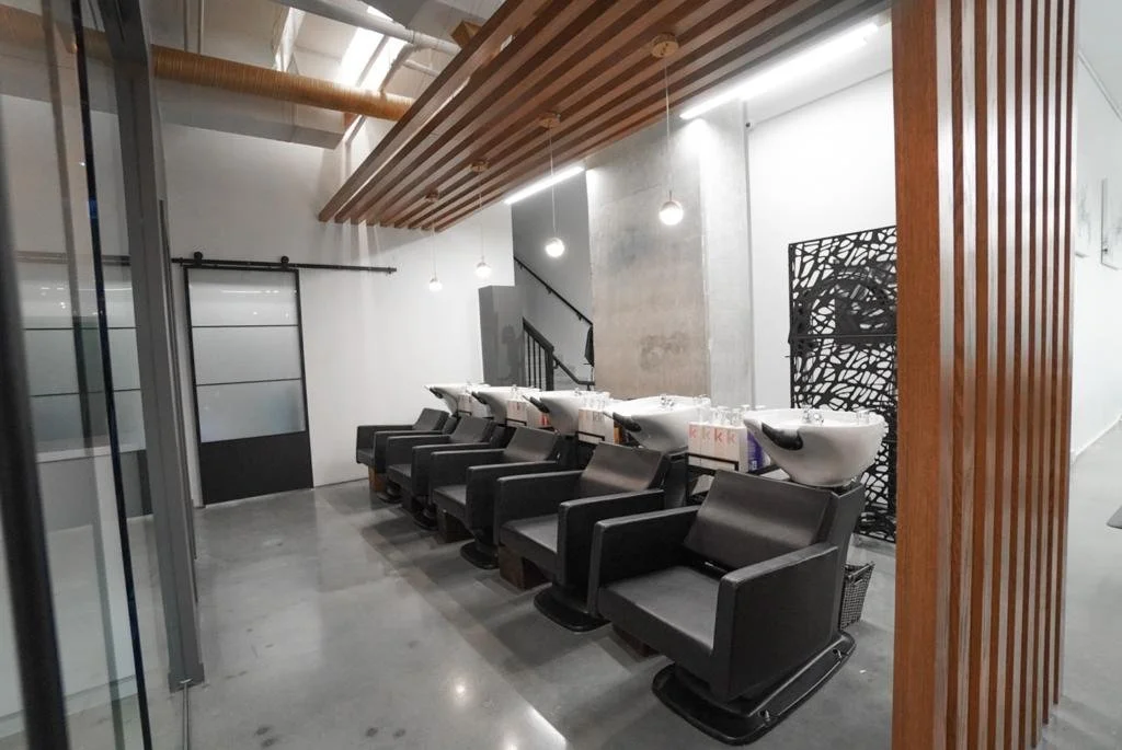 Hair salon with black styling chairs and washing basins, modern decor with wooden accents and pendant lighting.