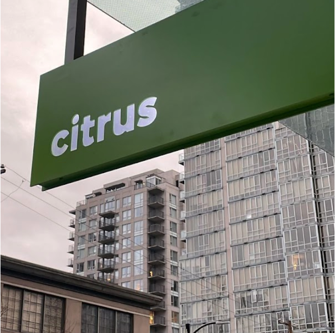 Green sign with white text that reads 'citrus,' hanging outside with modern high-rise buildings in the background.