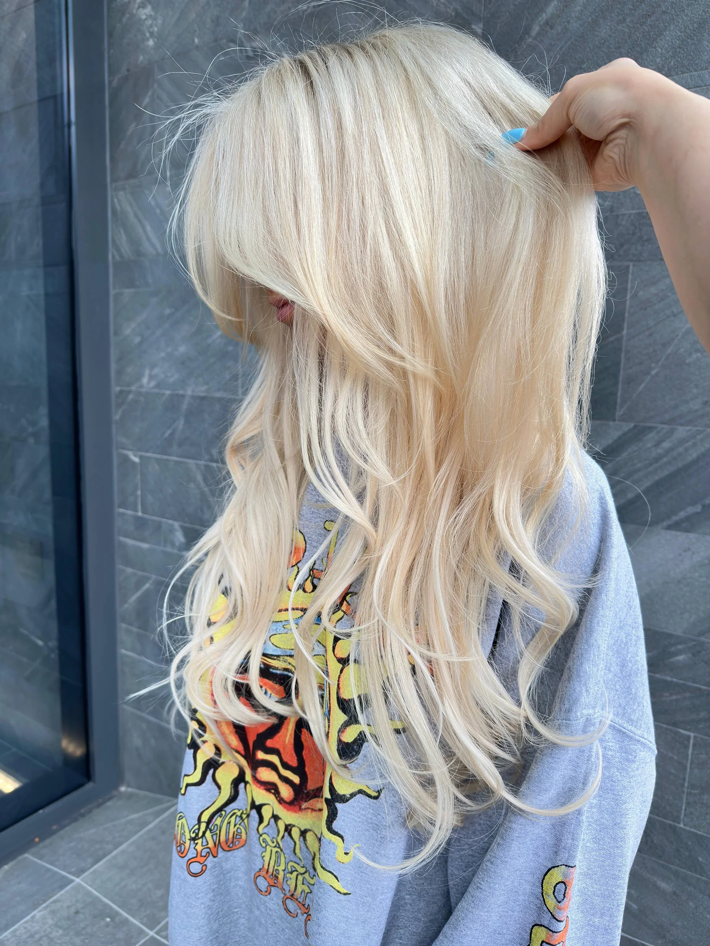 Person with long, wavy platinum blonde hair adjusting their hair in a casual setting with dark gray tiled wall.