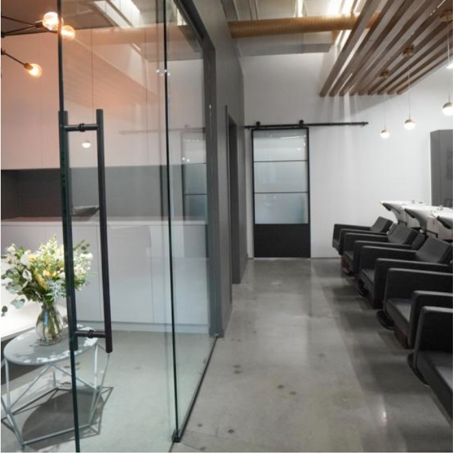 Modern hair salon with black styling chairs, washing stations, and a glass door, featuring a white wall and wooden ceiling accents.