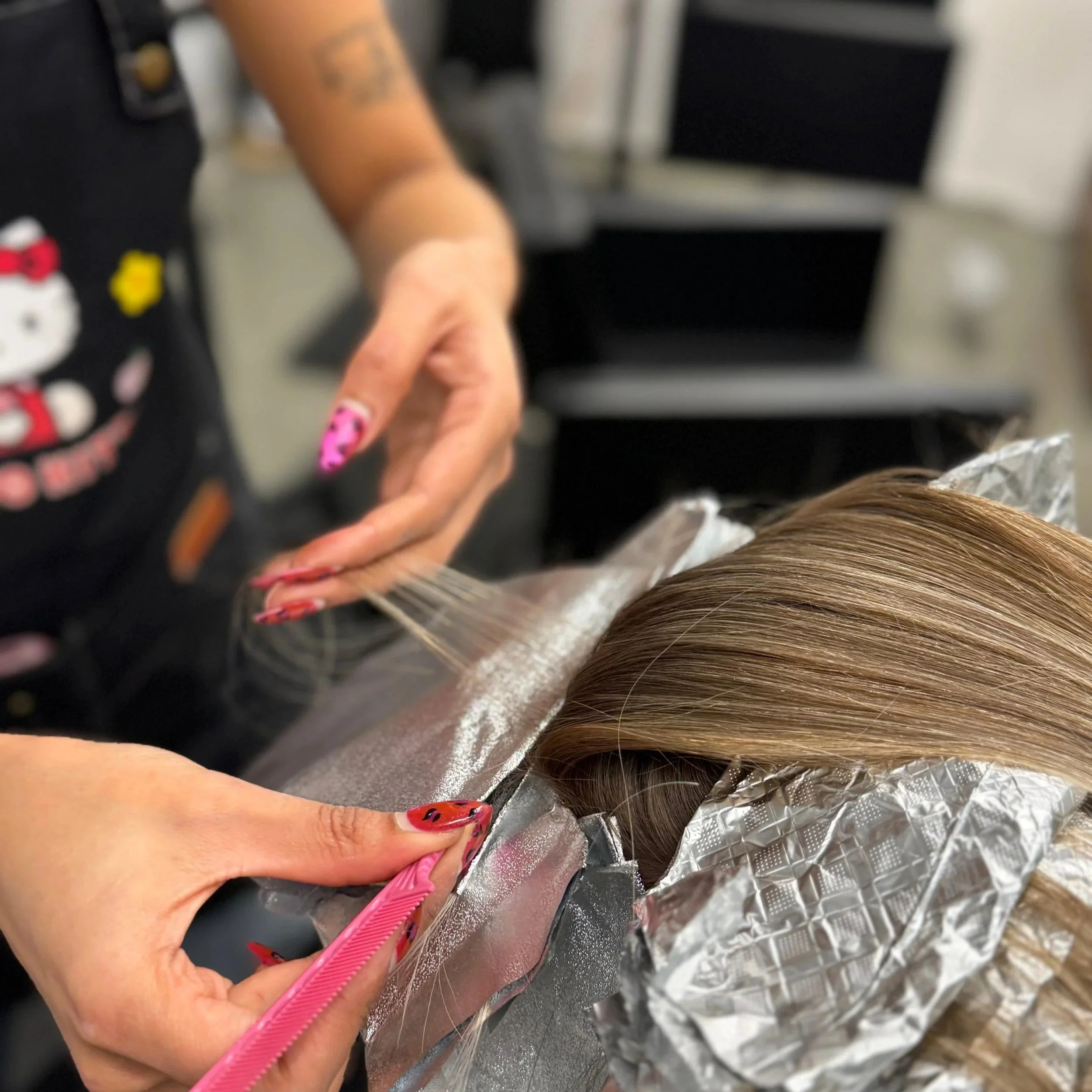 Hairdresser applying hair extensions or highlights to a client's hair using a pink comb, with the client lying back in a salon chair, hair sectioned and covered with foil.