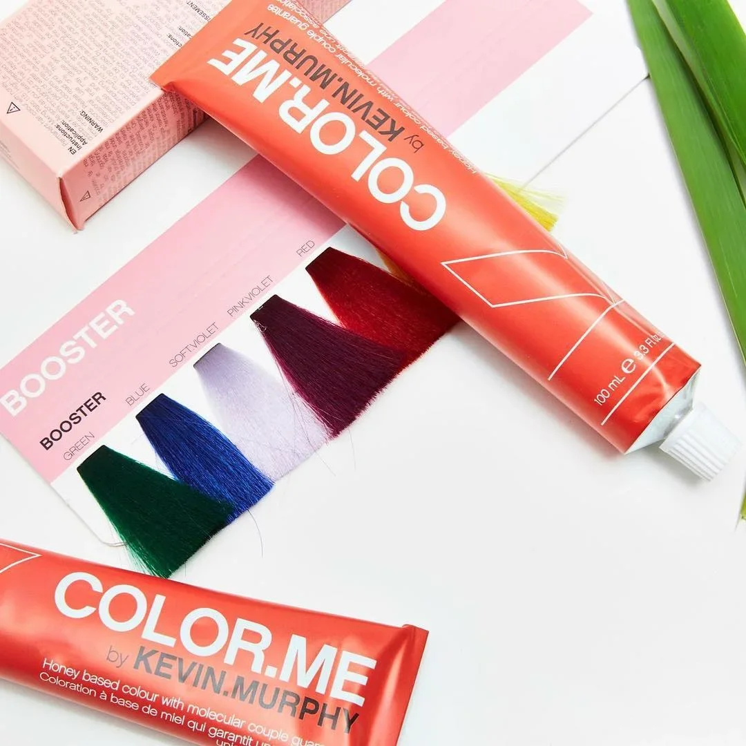 Hair color products including a coral-colored box labeled 'COLOR.ME by KEVIN.MURPHY', a tube, and a promotional card displaying a color palette with green, blue, soft violet, pink violet, and red swatches, on a white background with green leaves in the corner.