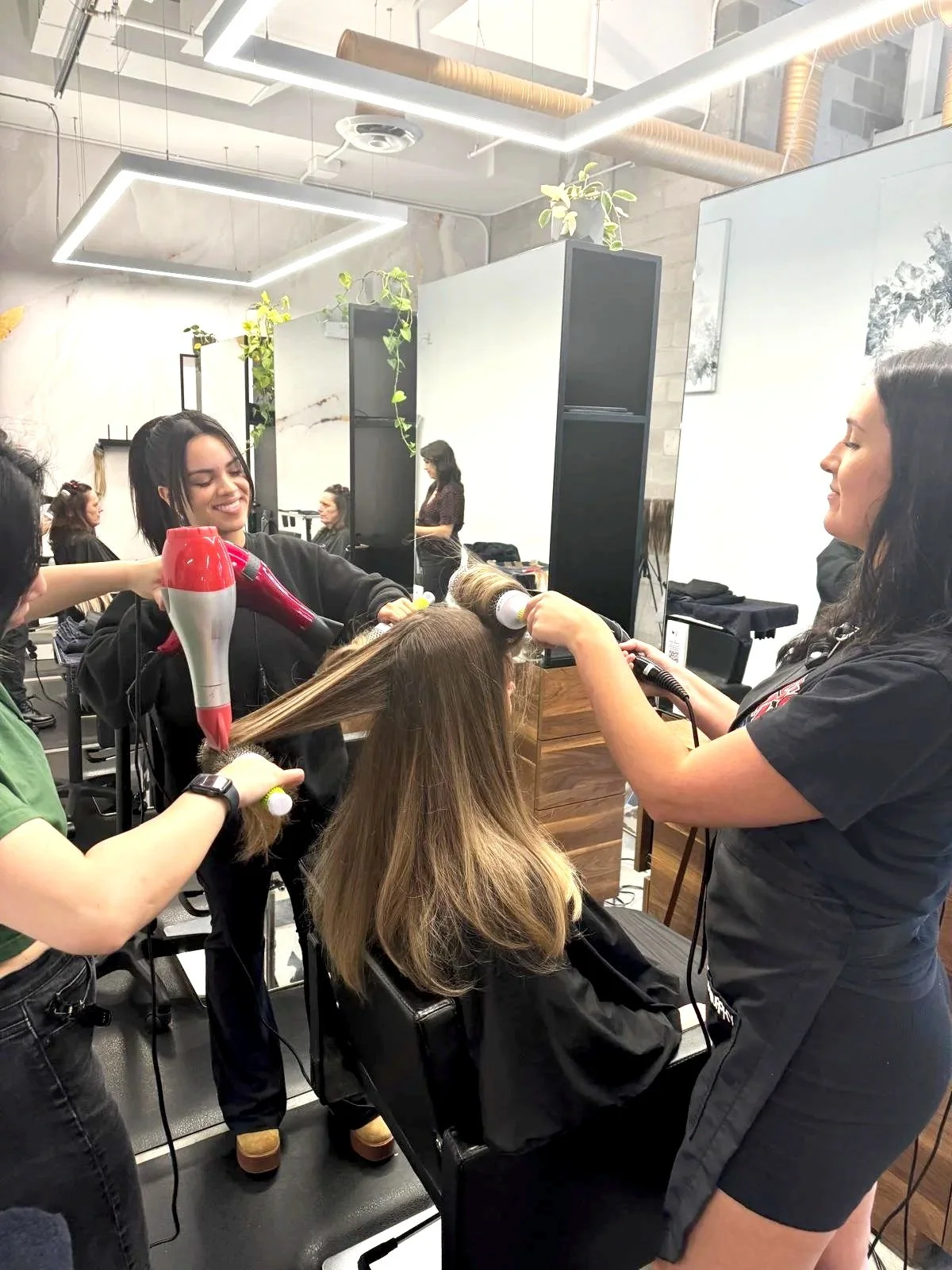 A woman with blonde hair getting her hair styled or dried by a hairstylist, smiling, in a modern hair salon with other clients and stylists in the background.