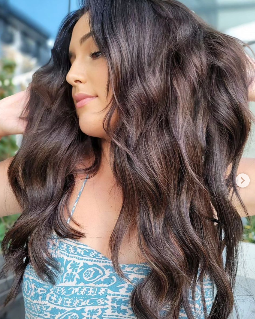 A woman with long, wavy, dark brown hair with subtle highlights, wearing a blue and white patterned top, with her eyes closed and a gentle smile.