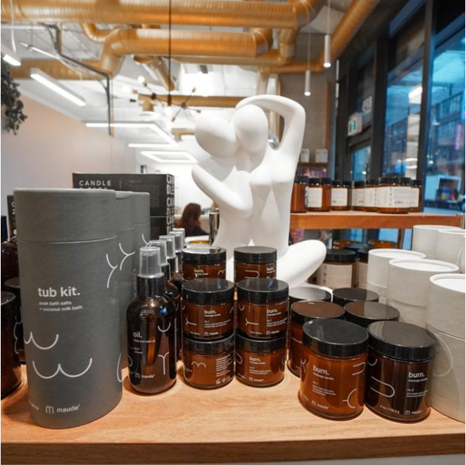 Display of bath and body products including a mock tub kit, body creams labeled 'burn.', and small bottles, with abstract white sculpture of a seated figure and store shelves in the background.