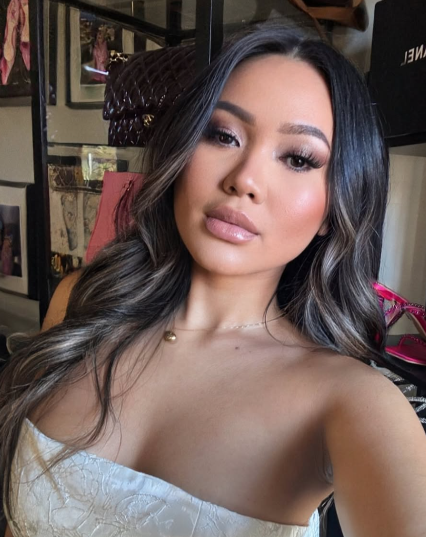 Close-up of a woman with long, wavy hair, wearing makeup and a white off-shoulder top, taking a selfie indoors.