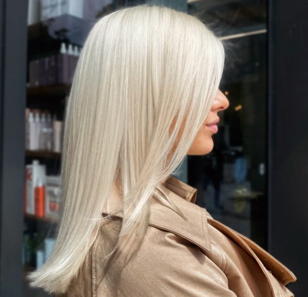 Profile side view of a woman with long, straight platinum blonde hair, wearing a beige trench coat, standing outdoors in front of a glass door.