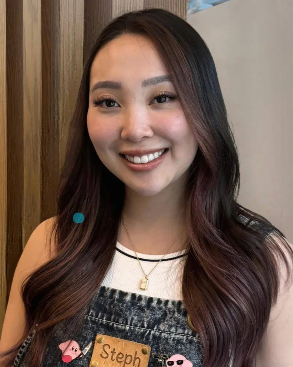 A smiling young woman with long wavy dark hair with reddish highlights, wearing a black and white top and a denim overall with pins, one of which has the name 'Steph'. She is also wearing a gold necklace with a small pendant.