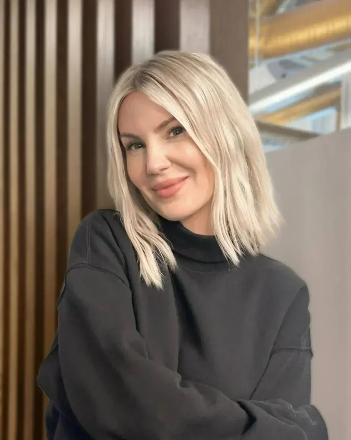 A young woman with shoulder-length blonde hair, wearing a black top, sitting indoors with a smile.