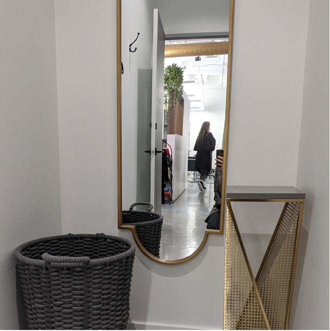 A tall mirror with a gold frame leaning against a white wall. In front of the mirror, there are two woven baskets, one small and one large, both dark gray. To the right of the mirror, there is a small gold and glass side table. The reflection in the 