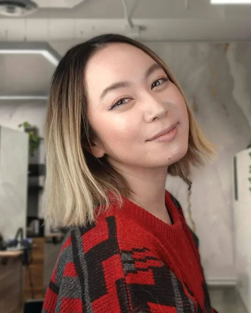 A young woman with shoulder-length ombré hair, wearing a red and black patterned sweater, smiling gently at the camera in an indoor space.