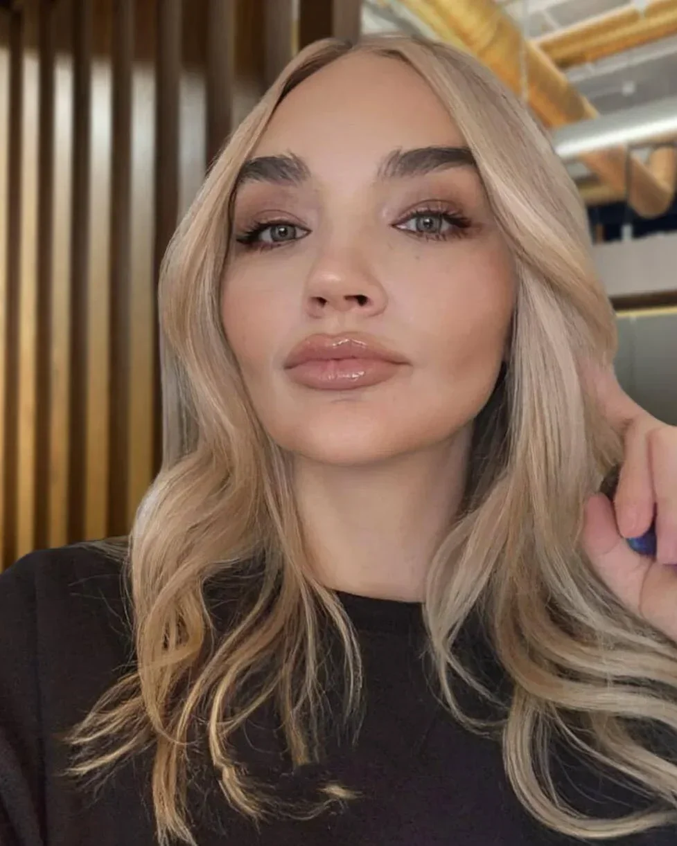 Close-up portrait of a young woman with wavy blonde hair, wearing a black top, in an indoor setting with wooden and metallic decor.