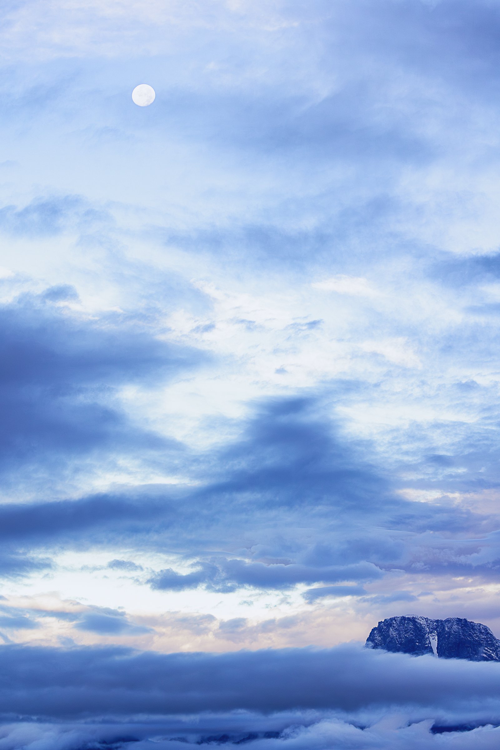 High above a sea of drifting clouds, a luminous moon watches over a solitary mountain summit emerging from the mist. Soft layers of blue and lavender stretch across the sky, creating an atmosphere of tranquility and mystery. This ethereal landscape c