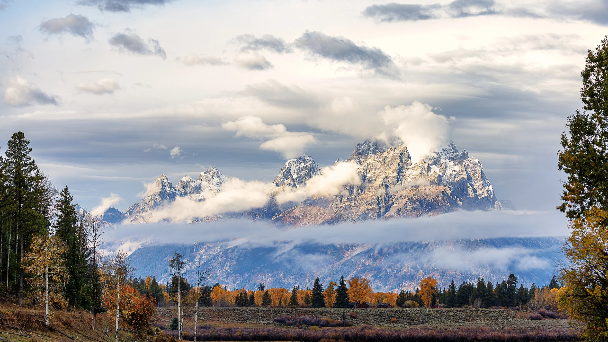 The towering peaks of the Teton Range emerge through drifting clouds as autumn paints the valley below in brilliant shades of gold and amber. Sunlight dances across the rugged, snow-dusted summits, while ribbons of mist weave through the mountainside