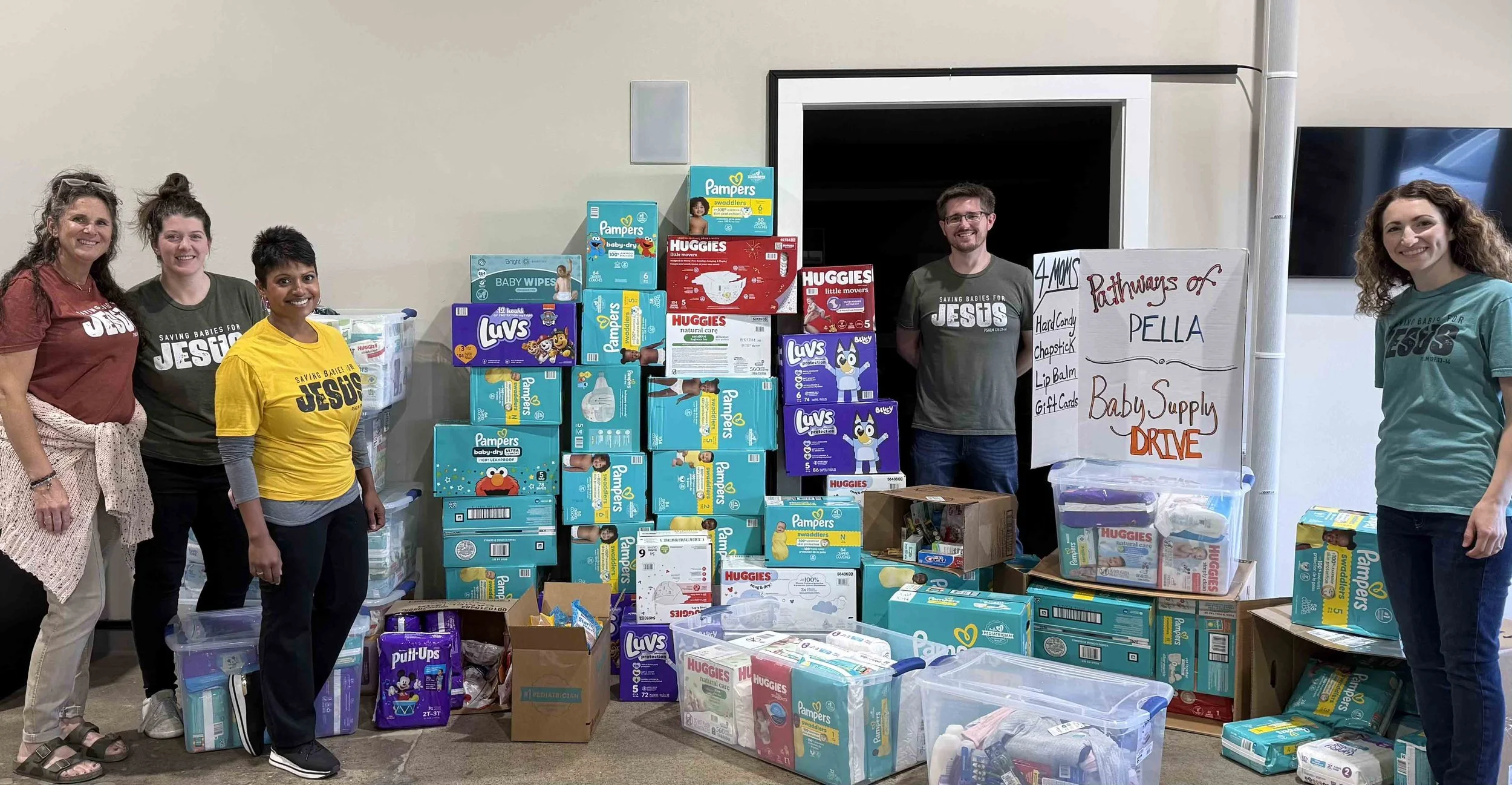 Five women standing next to a large collection of diaper boxes, baby supplies, and donation boxes, with a whiteboard listing a diaper drive and baby supply list.