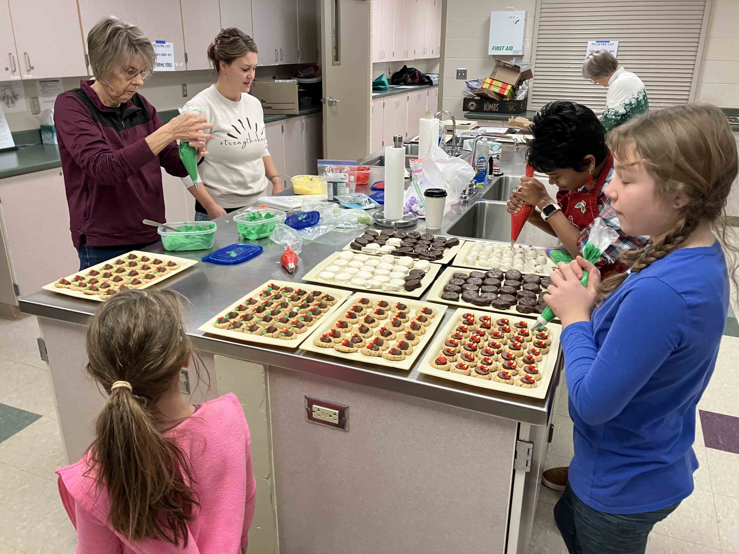 Children and adults decorating cookies with frosting and sprinkles in a kitchen.