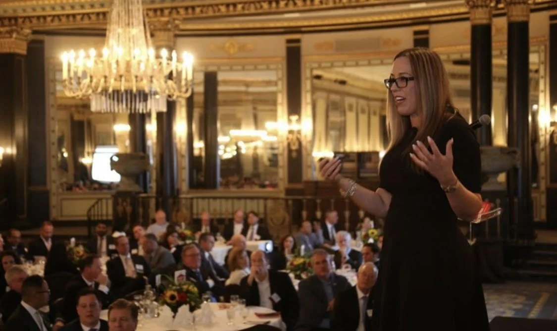 A woman with long blonde hair, glasses, and a black dress speaking at a formal event in a grand ballroom with chandeliers and many seated guests.