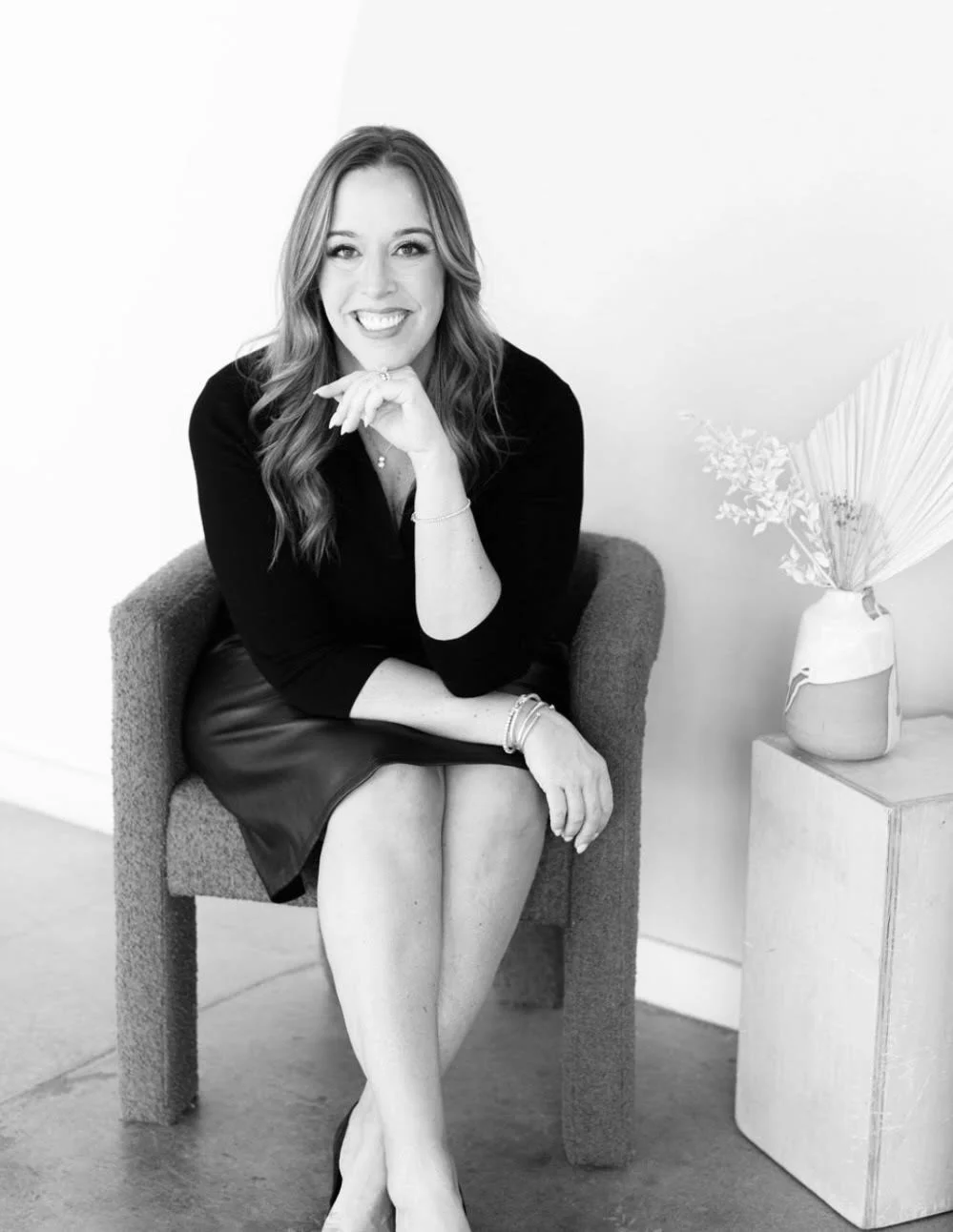 A woman with wavy hair sitting on a chair, smiling, wearing a black top and a leather skirt, with her legs crossed and her left hand resting on her chin, in a minimalist room with a side table and a vase with dried leaves.