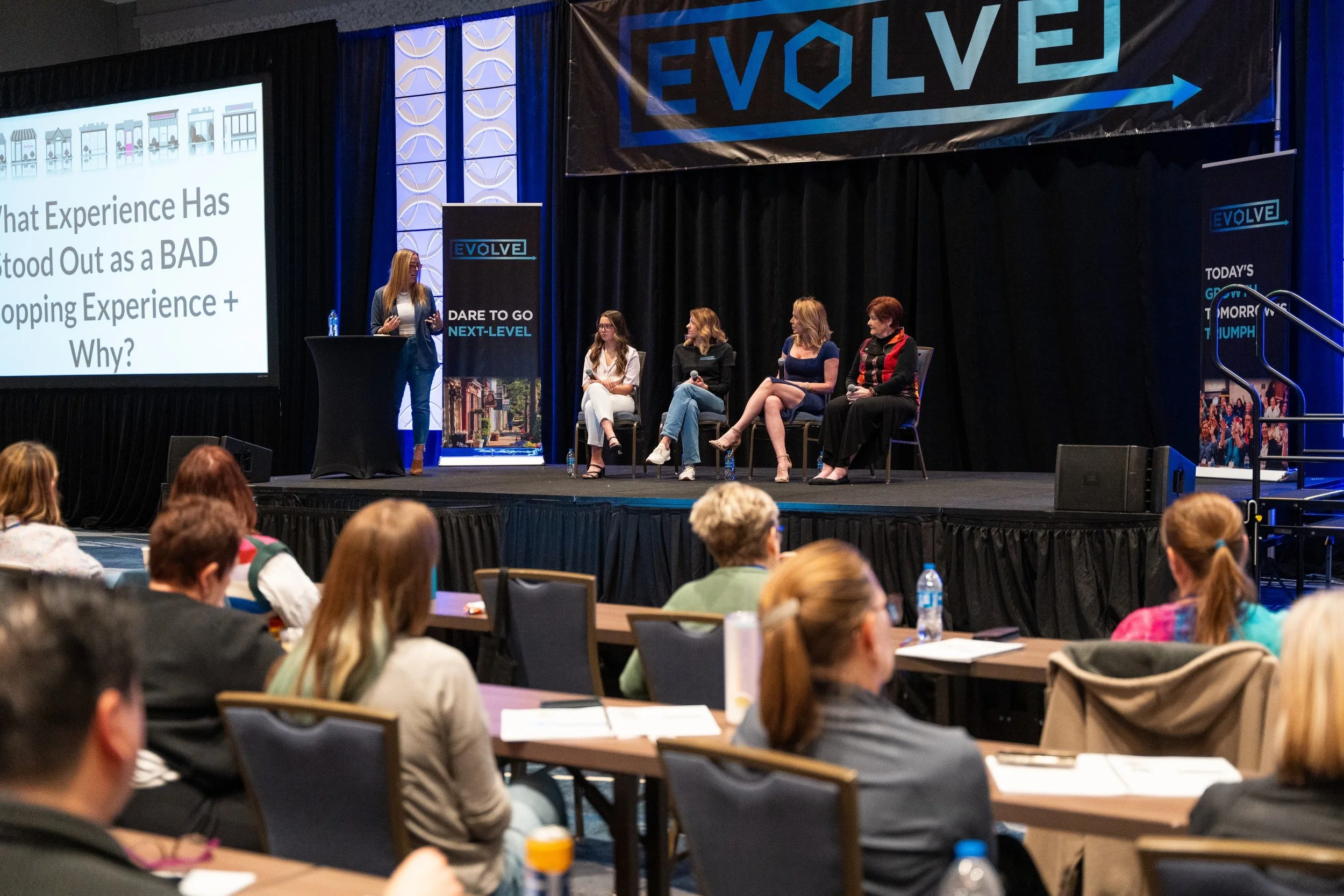 A conference panel discussion on stage with four women seated, one standing woman giving a presentation, and audience members listening in a large room.
