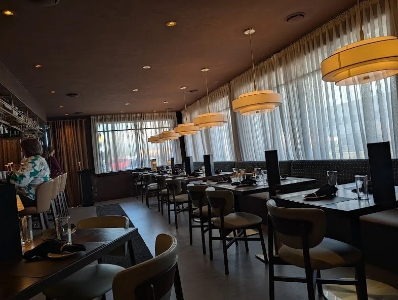 An empty restaurant with modern decor, hanging pendant lights, large windows with sheer curtains, and a few patrons at the bar.