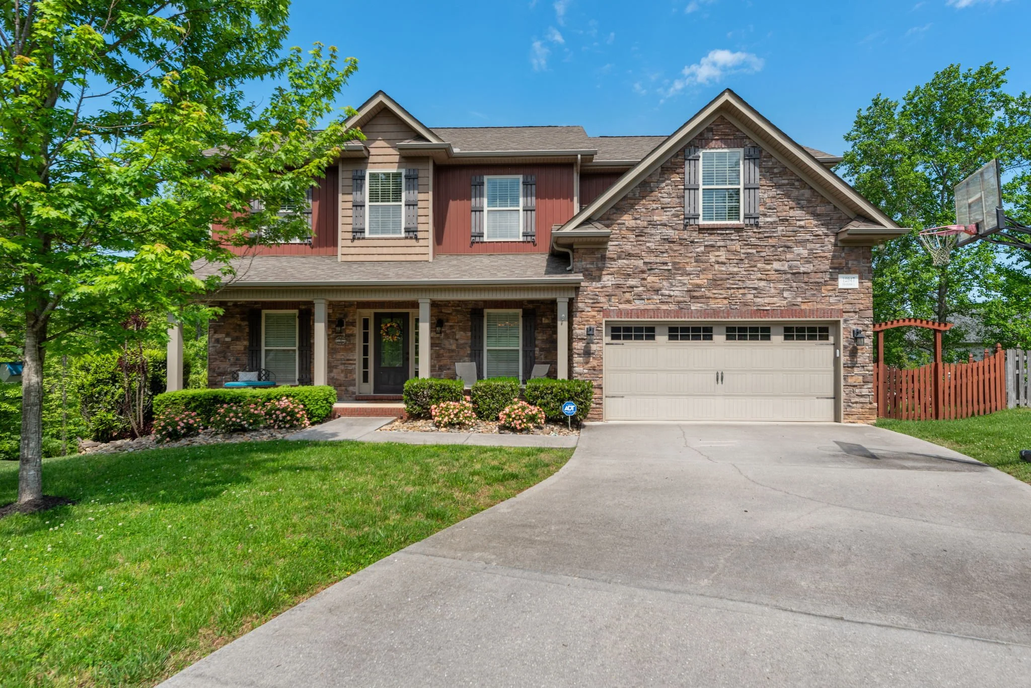 A two-story house with a combination of brick and wood siding facade, a green lawn, trees, and a concrete driveway leading to a two-car garage. There are shrubs and flowers in front, and a ball hoop on the right side of the driveway. The sky is blue with a few clouds.
