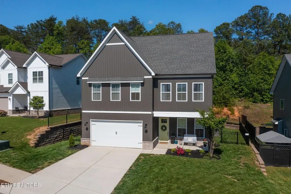 A modern two-story house with dark gray siding, a white garage door, and a small front porch with a white bench and houseplants. The house is surrounded by a green lawn, small trees, and other similar houses in the background, with a clear blue sky overhead.