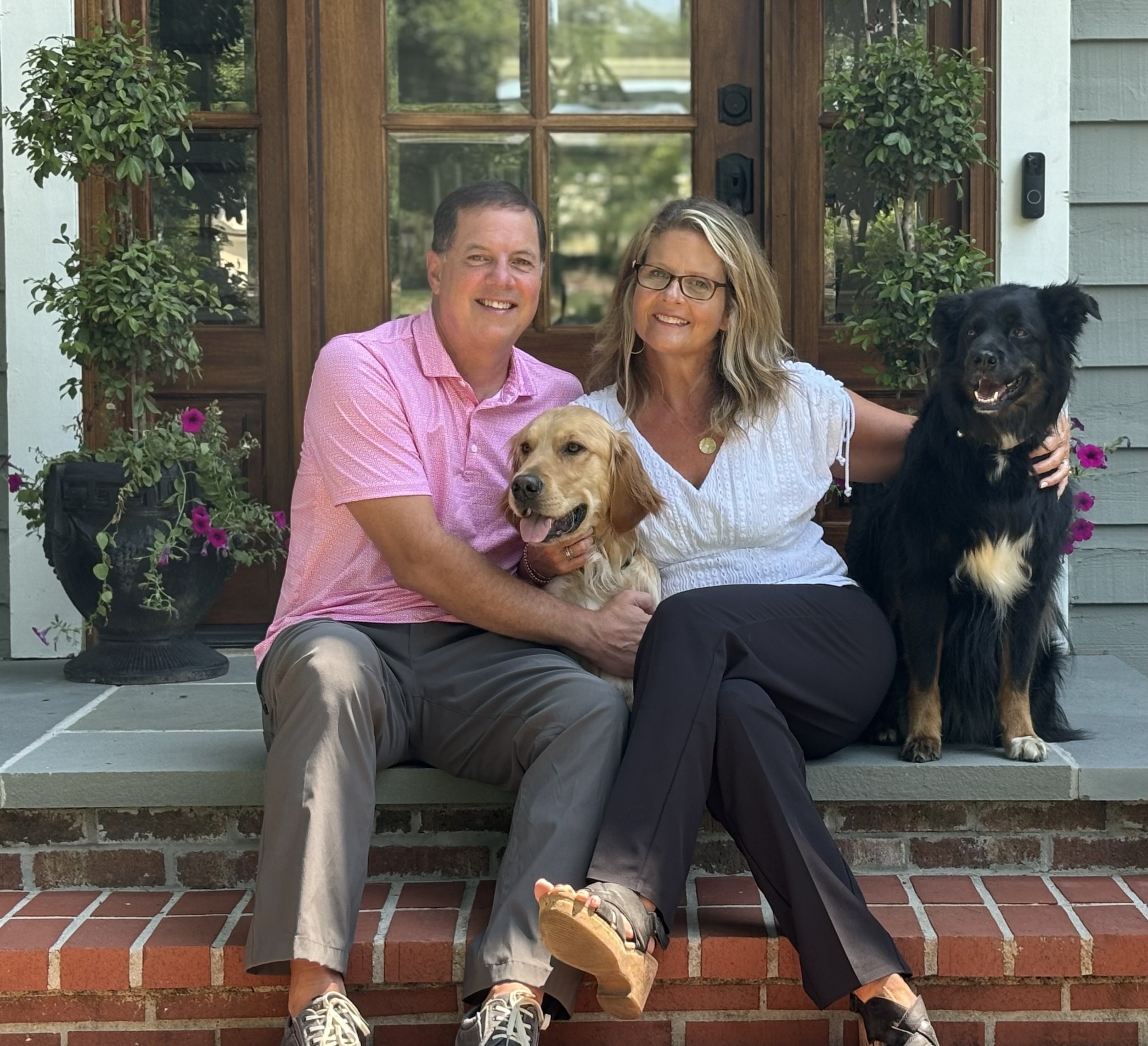 A smiling couple sitting on the porch steps with two dogs, one Golden Retriever and one black dog, in front of a house with a wooden door and potted plants.