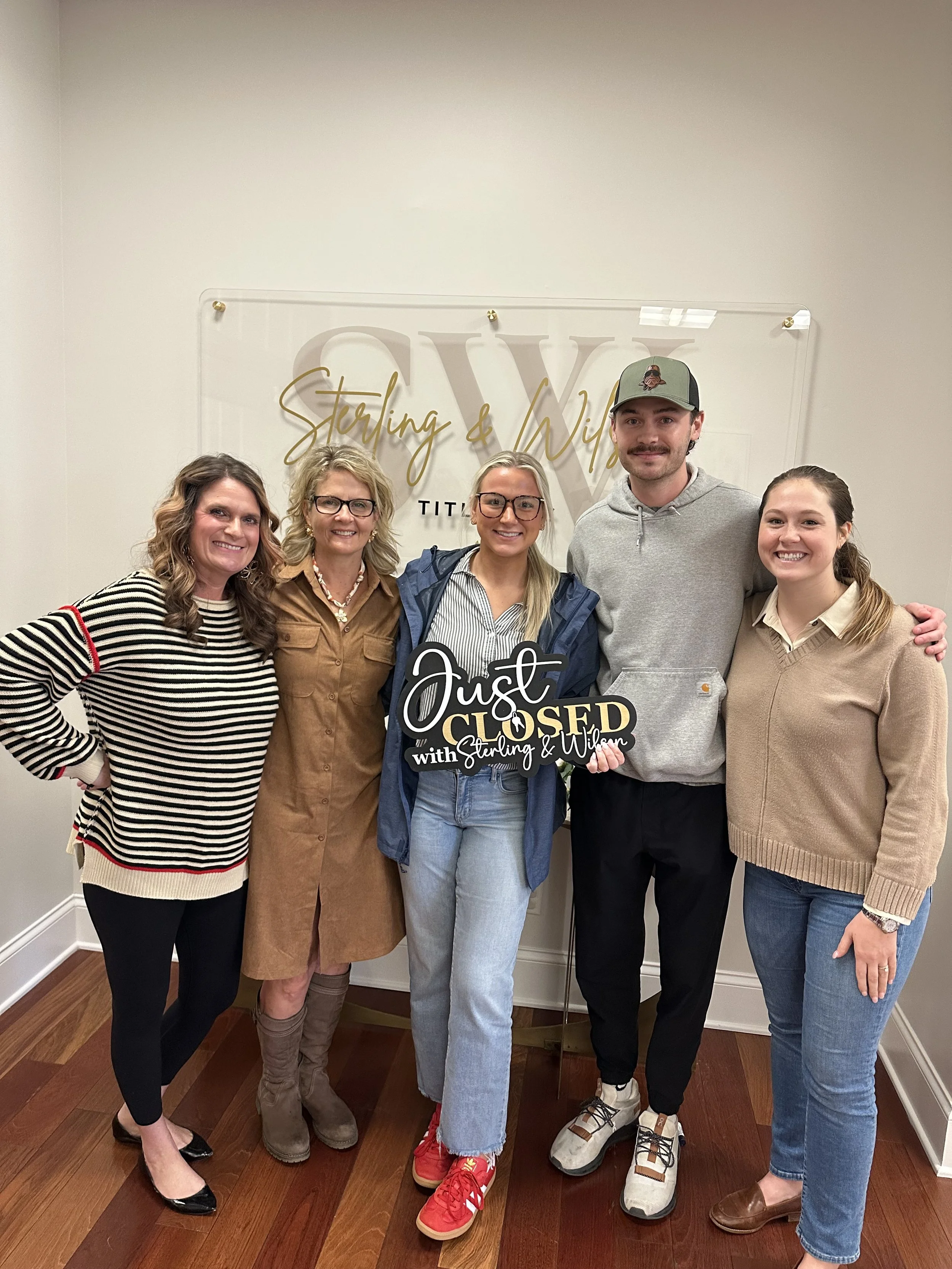 Group of five people standing together indoors, smiling, in front of a wall with a sign that reads 'Sterling & Wilson'. Two women are on the left and right edges, and in the middle are a woman with glasses and blonde hair, a man with a cap, and a woman with glasses and blonde hair. The woman in the middle is holding a sign that says 'Just Closed with Sterling & Wilson'.