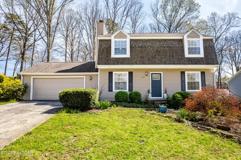 Front view of a two-story house with a beige exterior, gray shutters, a dark blue front door, a two-car garage, and a well-maintained lawn with shrubs and trees.