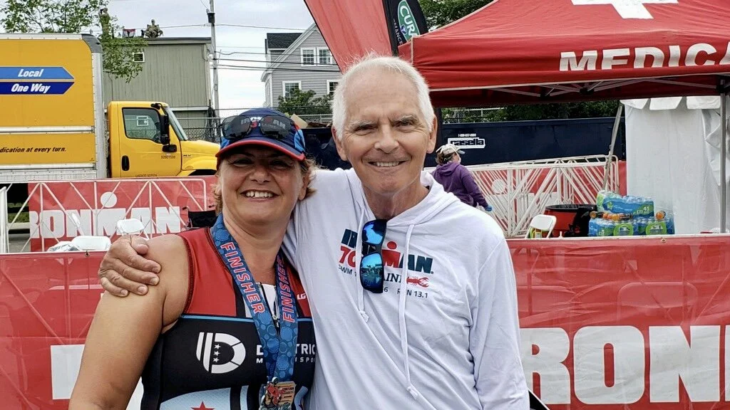A woman with a medal around her neck, dressed in athletic gear, standing next to a smiling older man in front of a red Ironman tent.